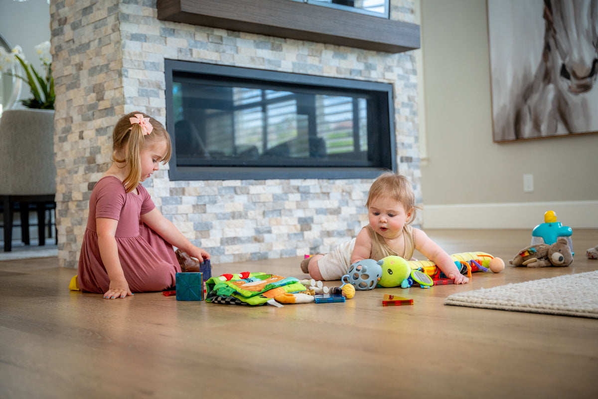 Two young children playing on a hardwood floor in front of a stone fireplace, surrounded by colorful toys and a soft fabric activity mat.