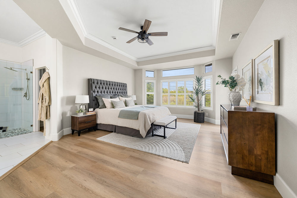 Light oak wood flooring in a spacious modern bedroom with a gray tufted bed, wooden dresser, and large windows.