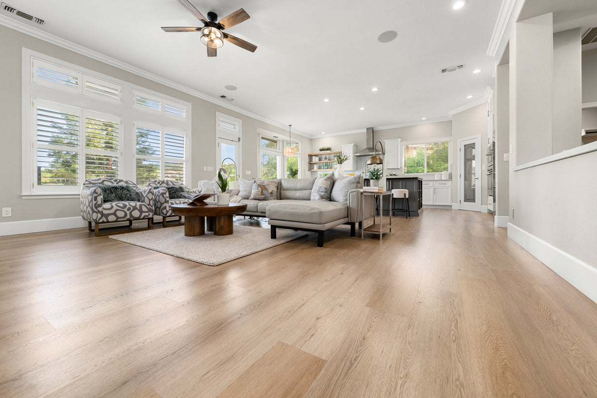 Light oak hardwood flooring in a bright open-concept living room with a gray sectional, patterned armchairs, and a kitchen.