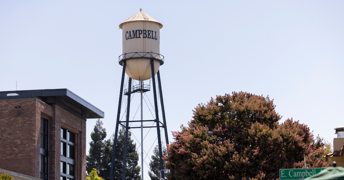 A tall Campbell water tower with a cream-colored tank and black metal support structure, photographed outdoors against a pale blue sky with trees and a brick building visible.