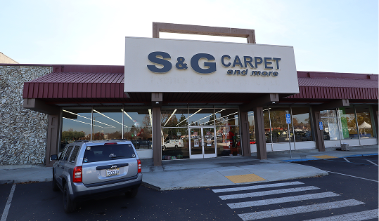 S&G Carpet and More storefront exterior with a large logo sign, glass entrance, and a silver SUV parked in front.