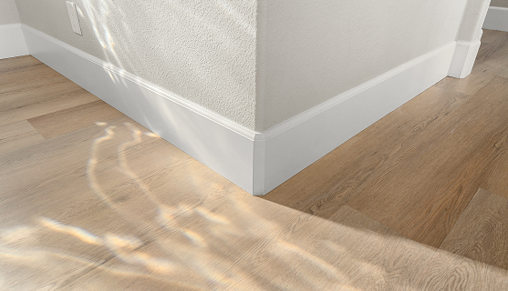 Light oak wood flooring and white baseboards installed along a wall corner with sunlight casting shadows.
