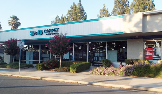 S&G Carpet storefront exterior with a white and teal building facade, large glass windows, and front landscaping.