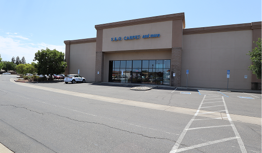 S&G Carpet storefront exterior with a beige facade, blue logo sign, glass entrance, and a wide paved parking lot.