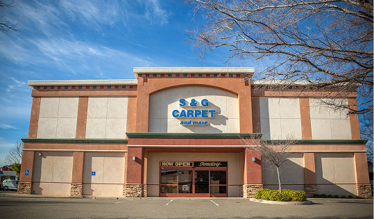 S&G Carpet and Tile storefront exterior under a blue sky, with bare tree branches and a paved parking lot.