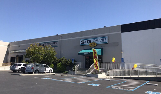 S&G Carpet storefront exterior with a gray building facade, green awning, entrance ramp, and parking lot.