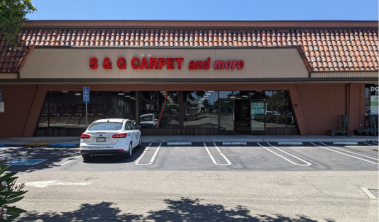 S&G Carpet and More storefront exterior with a red logo sign, glass entrance, and a white car parked in front.