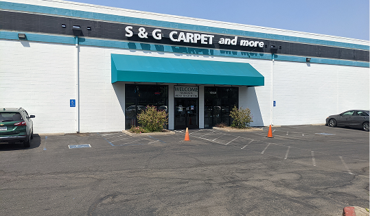 S&G Carpet and More storefront exterior with a white building facade, teal awning, and parking lot with orange cones.