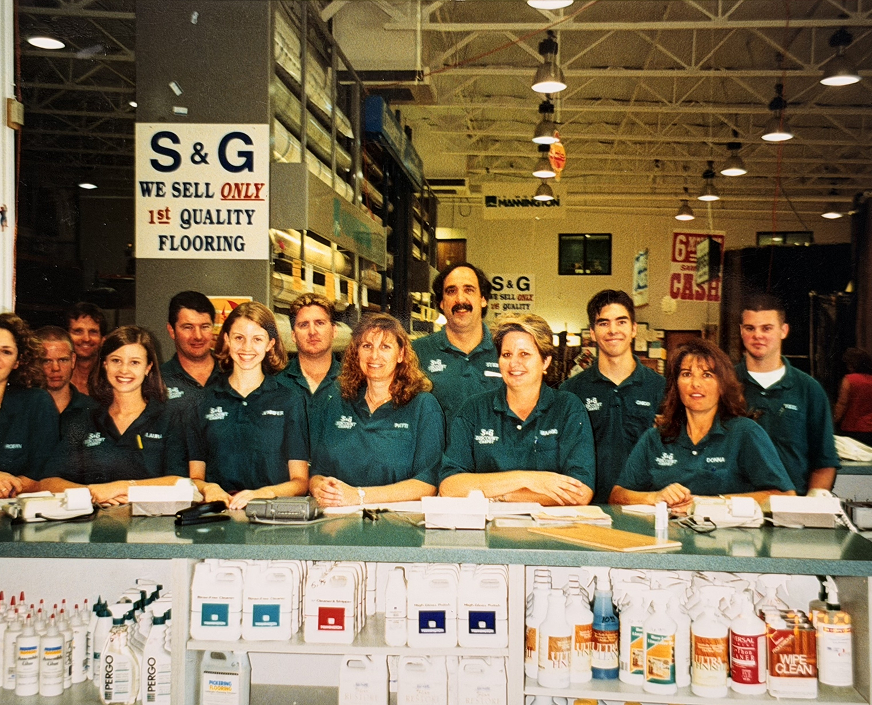 S&G Carpet employees in teal polos standing behind a store counter with a 1st Quality Flooring sign.