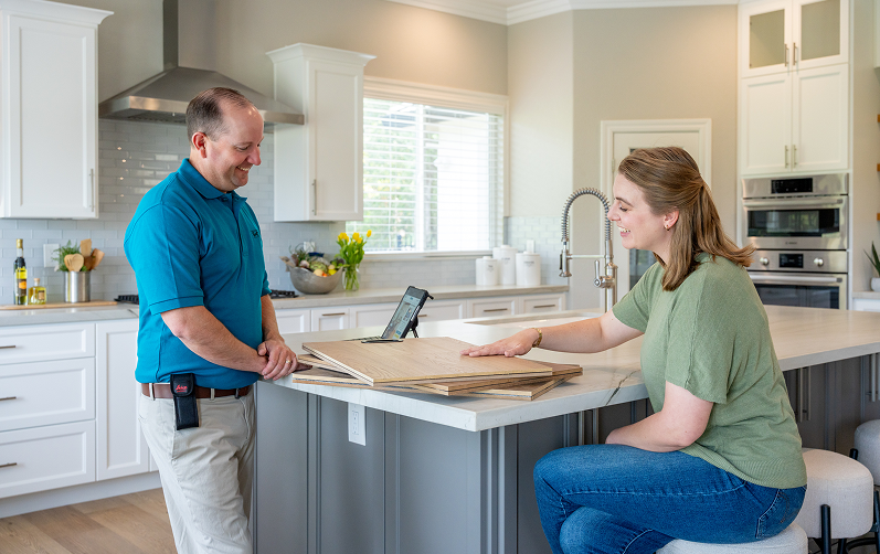 S&G Carpet estimator showing wood flooring samples to a homeowner on a gray island in a modern kitchen.