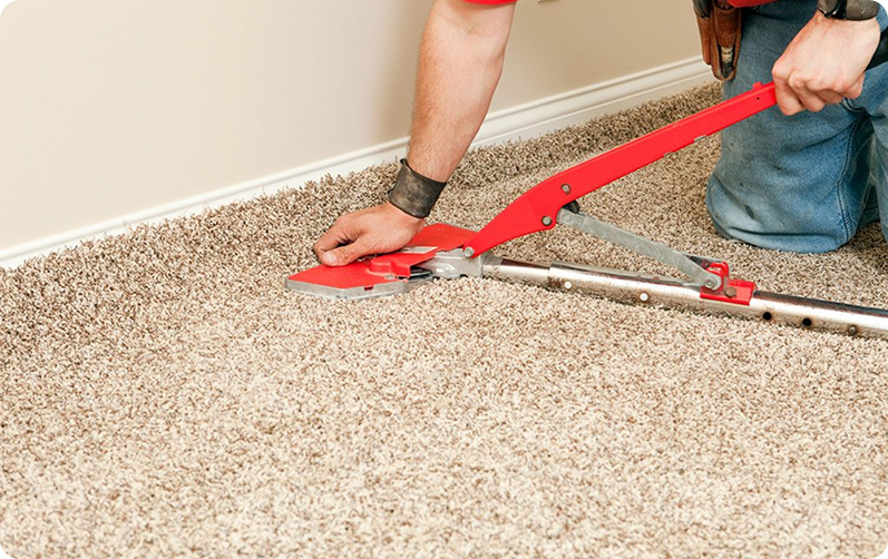 Installer using a red knee kicker tool to stretch and install beige carpet along a baseboard.