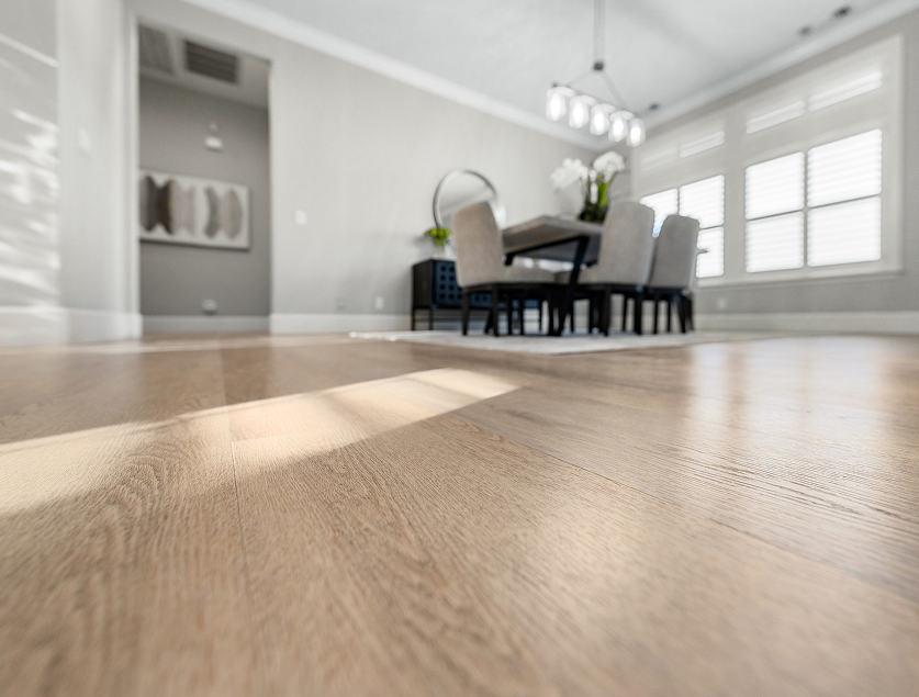Light oak hardwood flooring shown from a low angle with a blurred modern dining room and bright windows in the background.