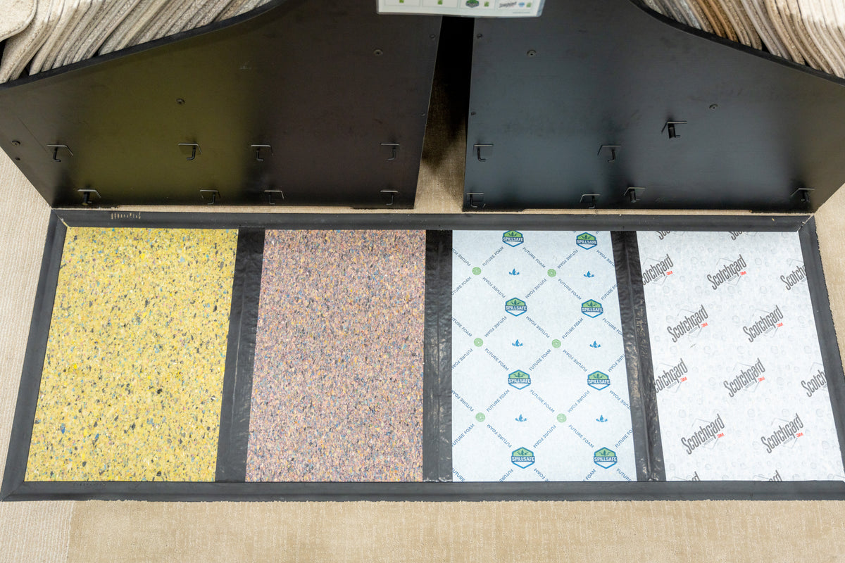 Four carpet padding samples in a black display tray on beige carpet, including speckled foam, SpillSafe, and Scotchgard pads.