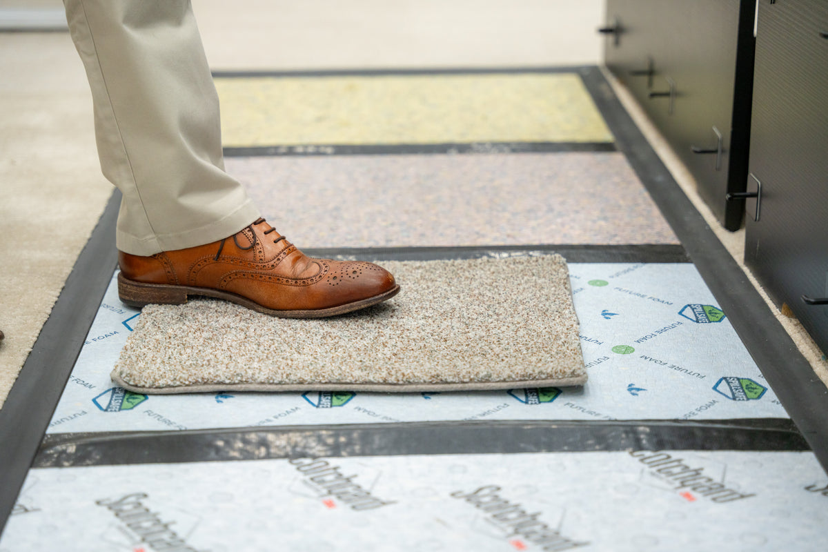 A person wearing beige trousers and a brown leather brogue shoe stepping onto a rectangular gray carpet sample, which is placed on top of branded carpet padding.