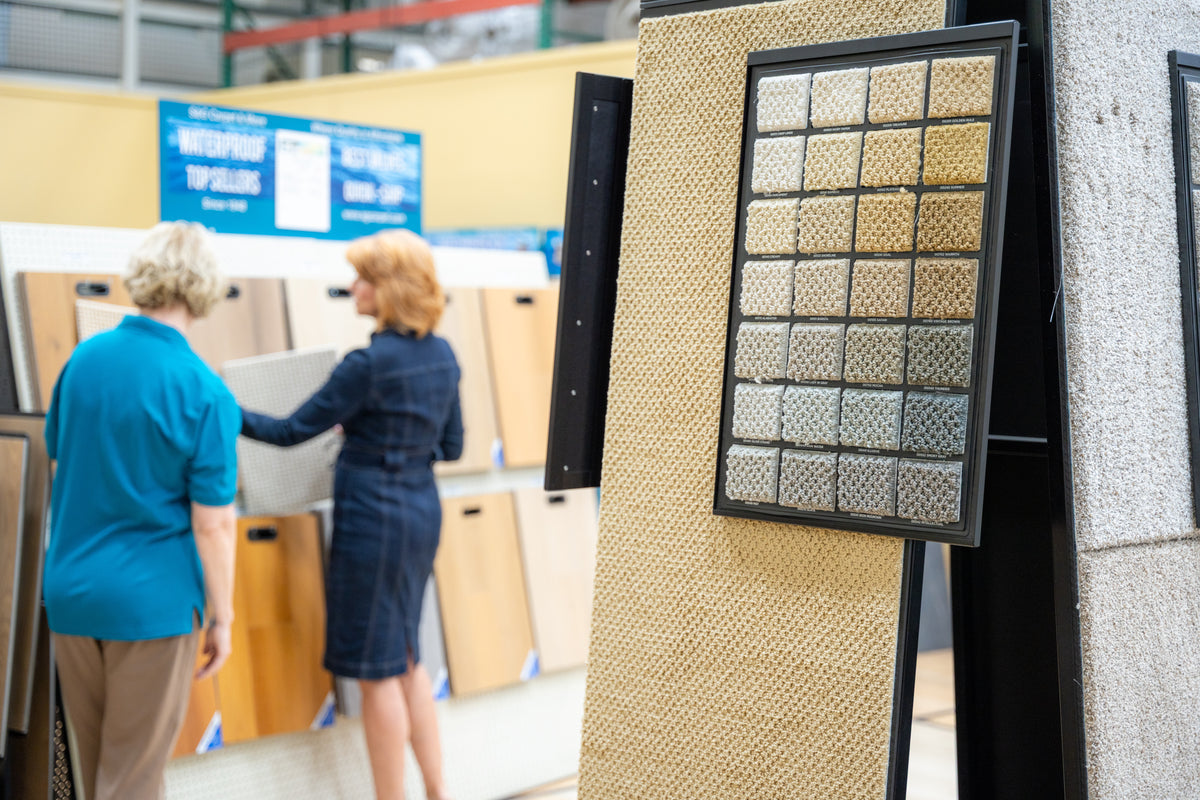 Board of neutral carpet color swatches in the S&G Carpet showroom as two shoppers browse samples behind it