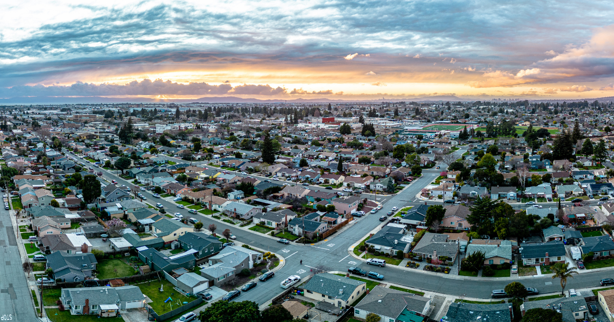 Aerial view of a San Leandro, suburban neighborhood with dense residential houses, streets, trees, and a dramatic cloudy sunset sky.