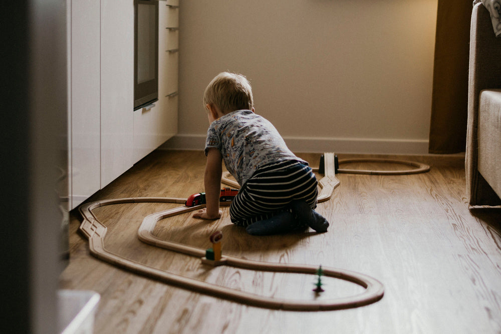 Toddler playing with a wooden train track on warm oak plank flooring in a living room