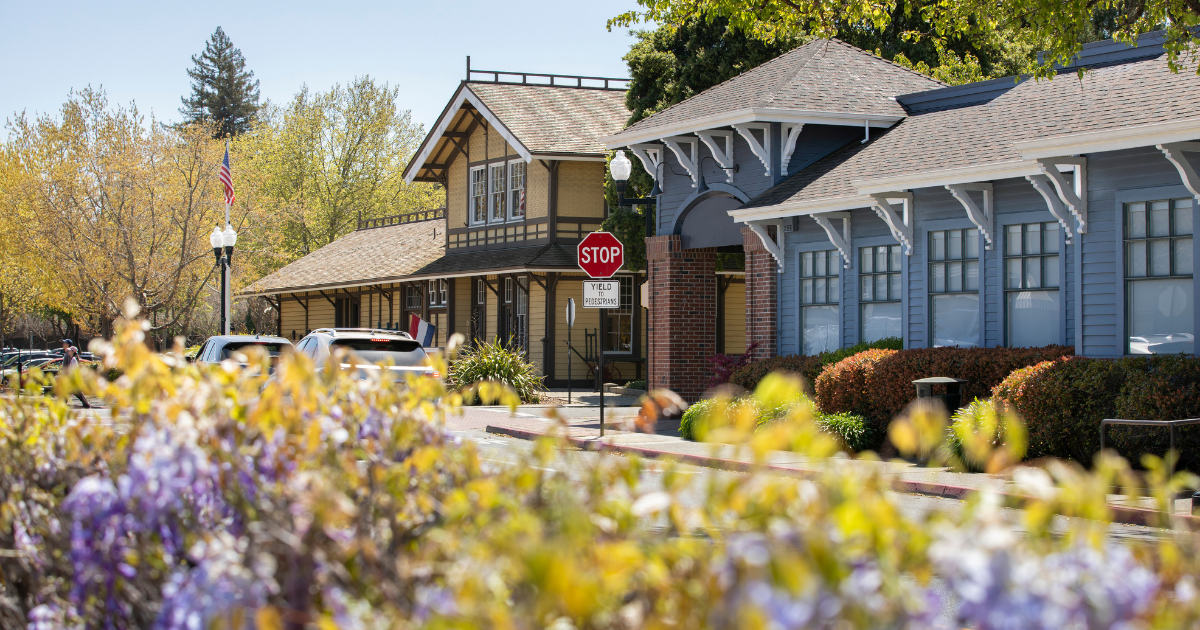 A scenic view of a historic-looking building complex with light blue and tan siding, set among green trees and blurred purple flowers in the foreground.