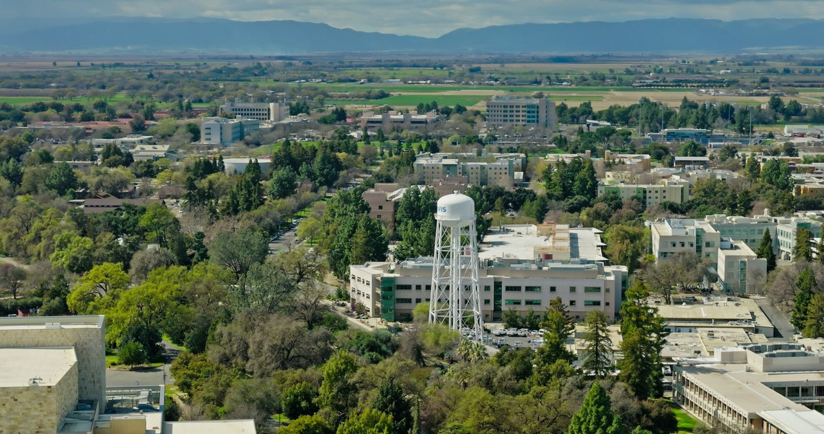 Aerial view of a campus-like urban landscape in Davis, California with buildings, trees, a white water tower, and distant mountains under a cloudy sky.