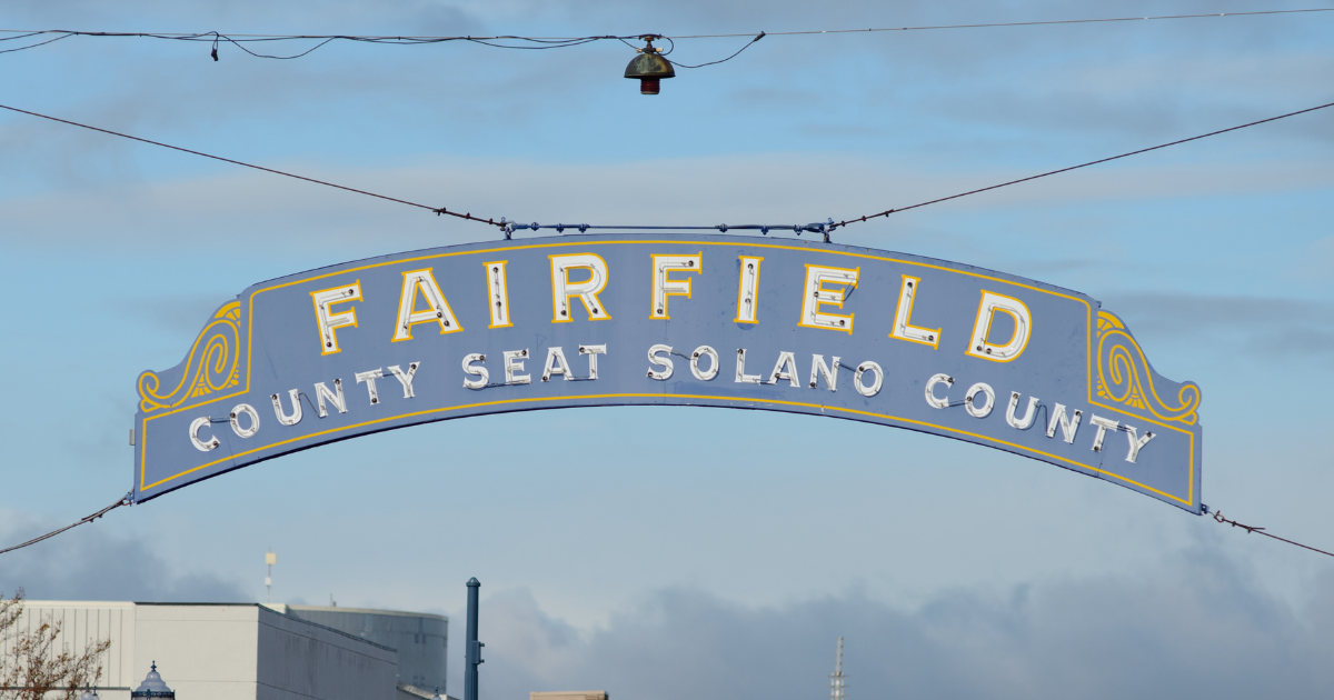 A large arched sign reading 'FAIRFIELD COUNTY SEAT SOLANO COUNTY' in gold and white lettering on a blue background, suspended outdoors against a cloudy sky.