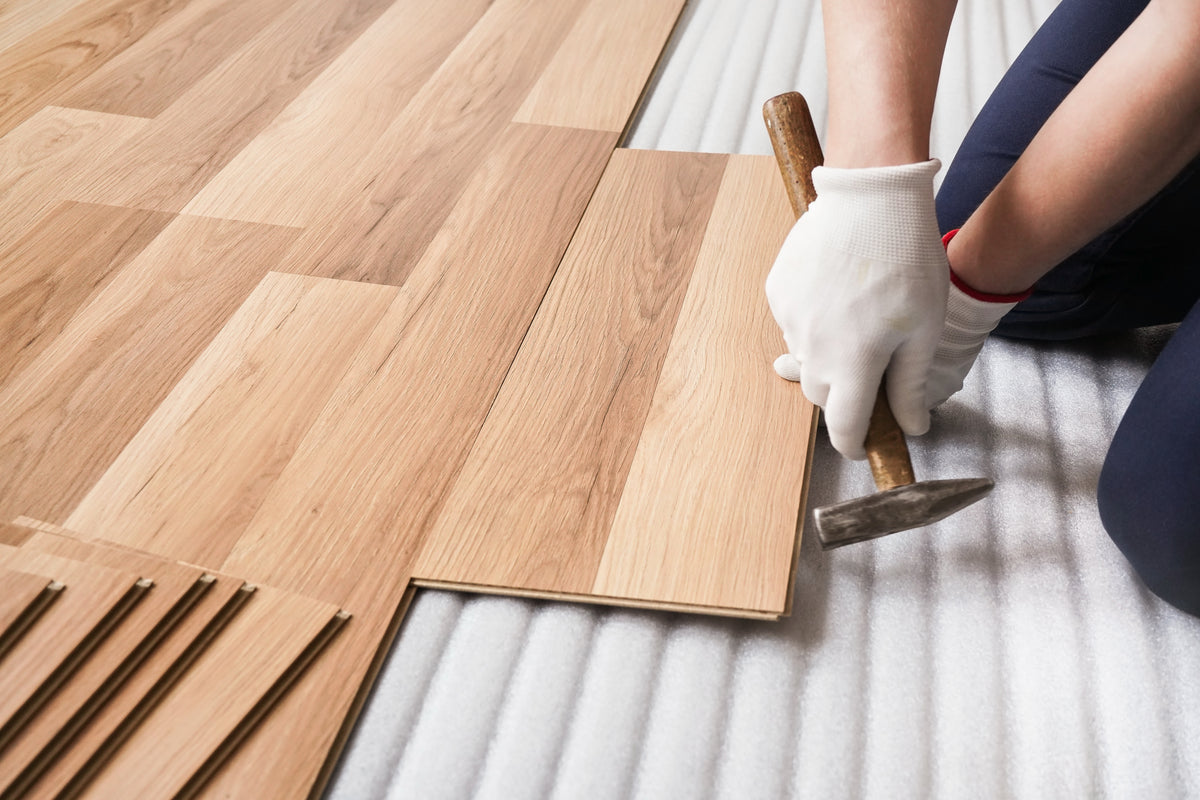 A close-up of a person wearing white work gloves installing light oak wood flooring planks, using a hammer to snap the planks into place on a white floor underlay.