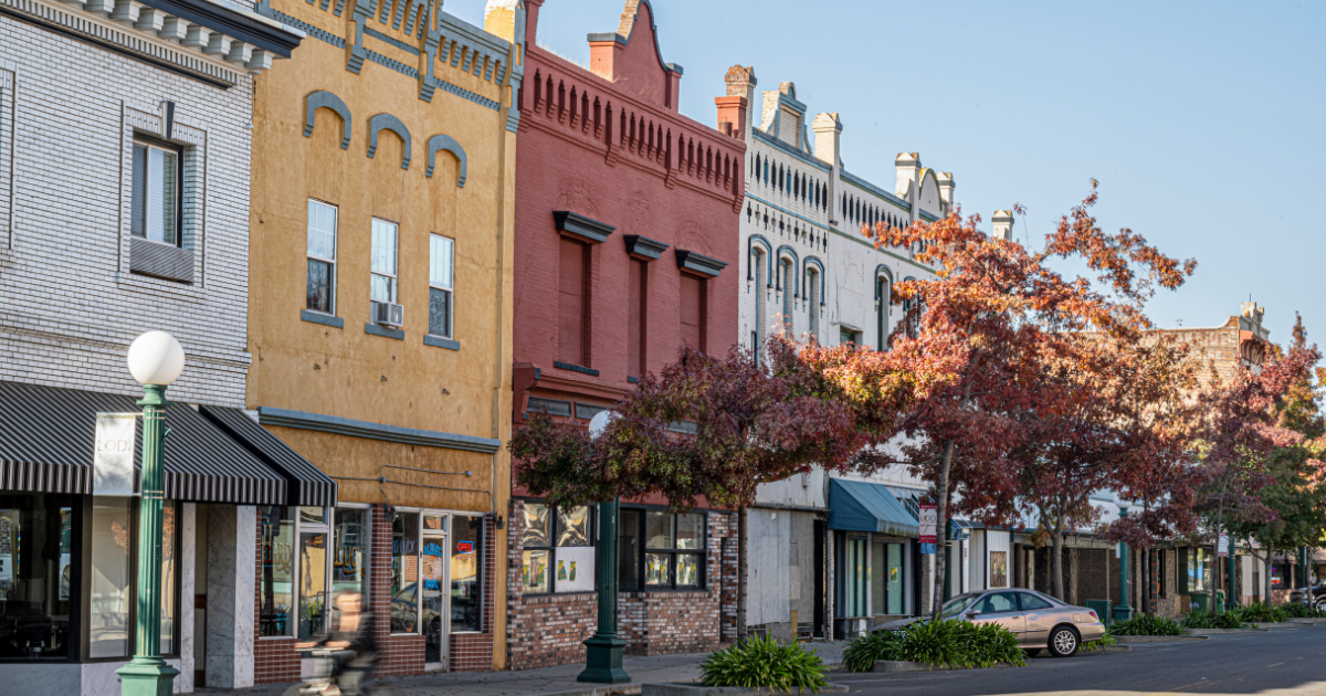 A scenic photograph of historic storefront buildings in Lodi, California, along a street, featuring brick and painted facades in beige, yellow, red, and white, with autumn trees in the foreground and a parked car on the street.