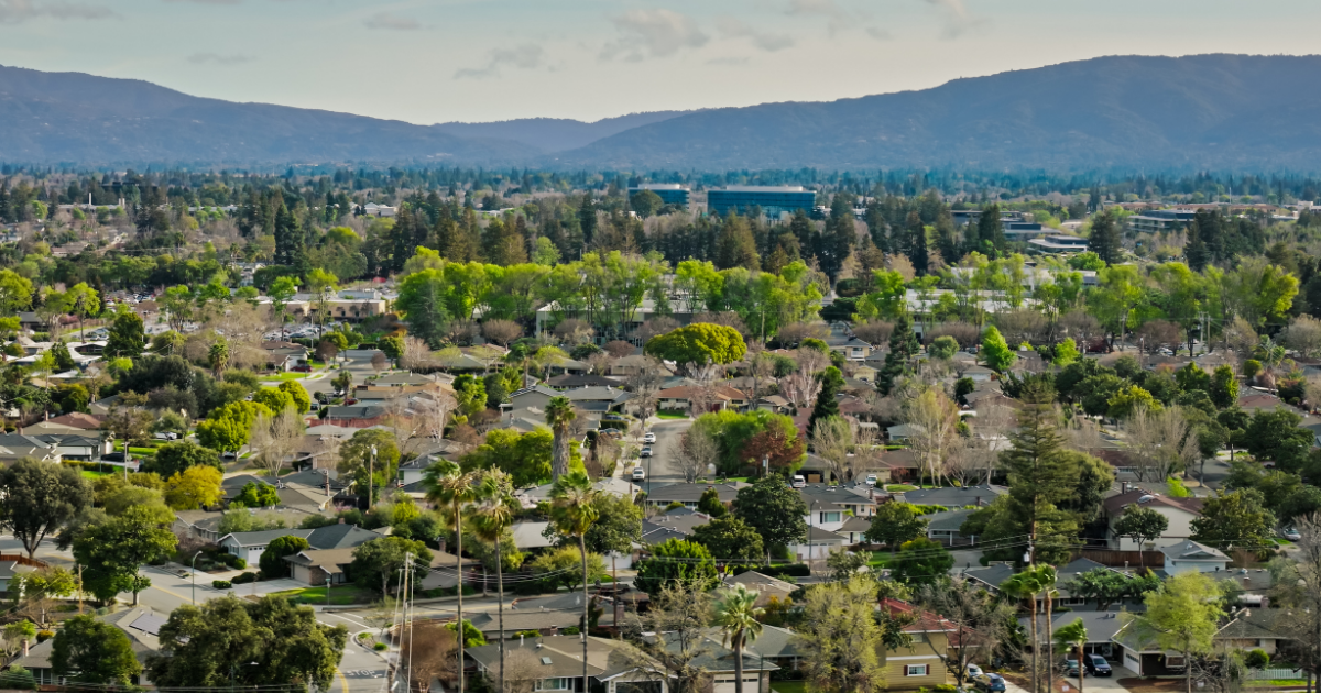 Aerial view of a Monte Vista neighborhood with dense trees, houses, and winding streets, with mountains in the background under a pale sky.
