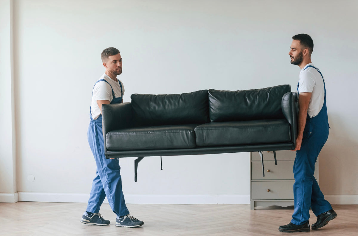 Two movers carrying a leather sofa across light herringbone wood flooring in an empty room