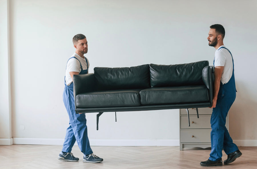 Two movers carrying a leather sofa across light herringbone wood flooring in an empty room