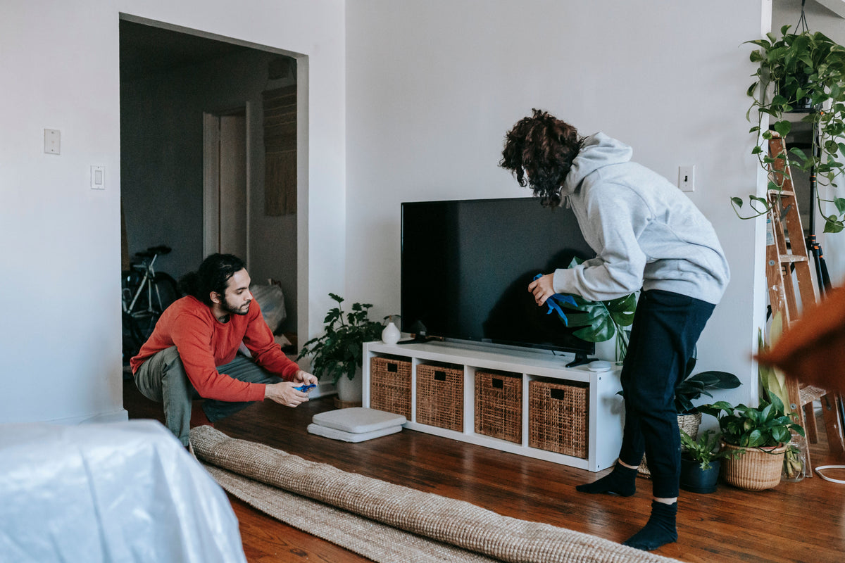 Two people setting up a TV console on dark hardwood flooring with a rug rolled back in a living room