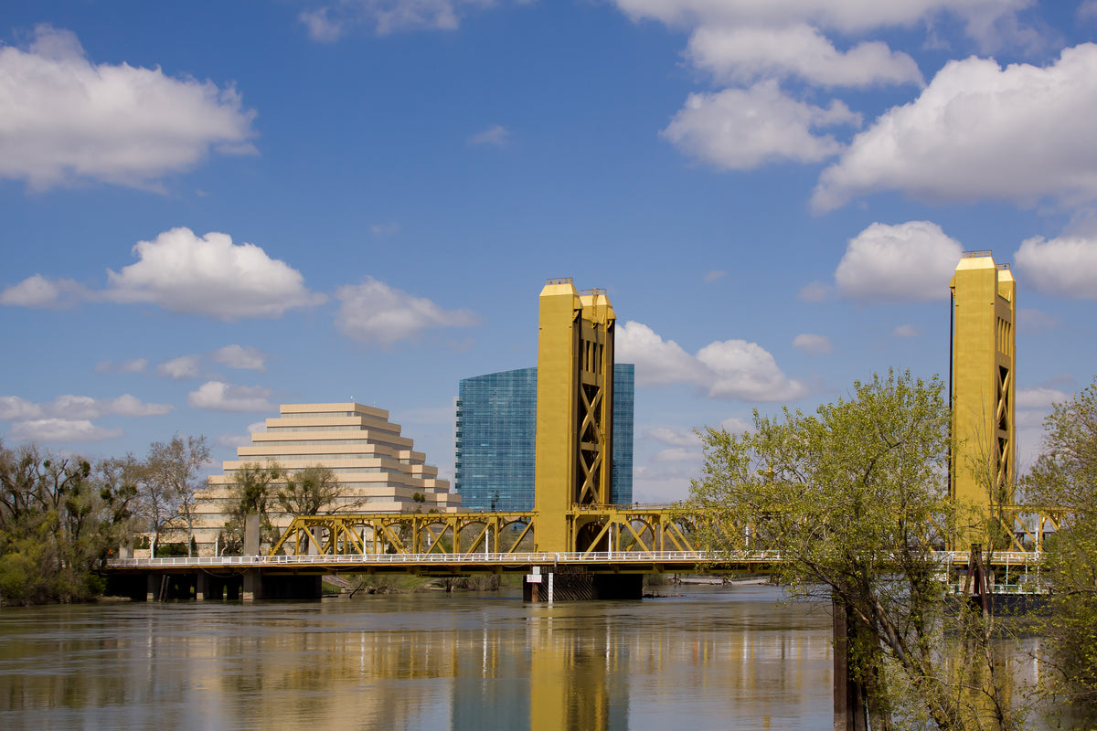 A scenic photograph of the bridge in West Sacramento, California, featuring its distinctive yellow towers and truss structure over the river, with the Pyramid and the blue glass building in the background under a blue sky with white clouds.
